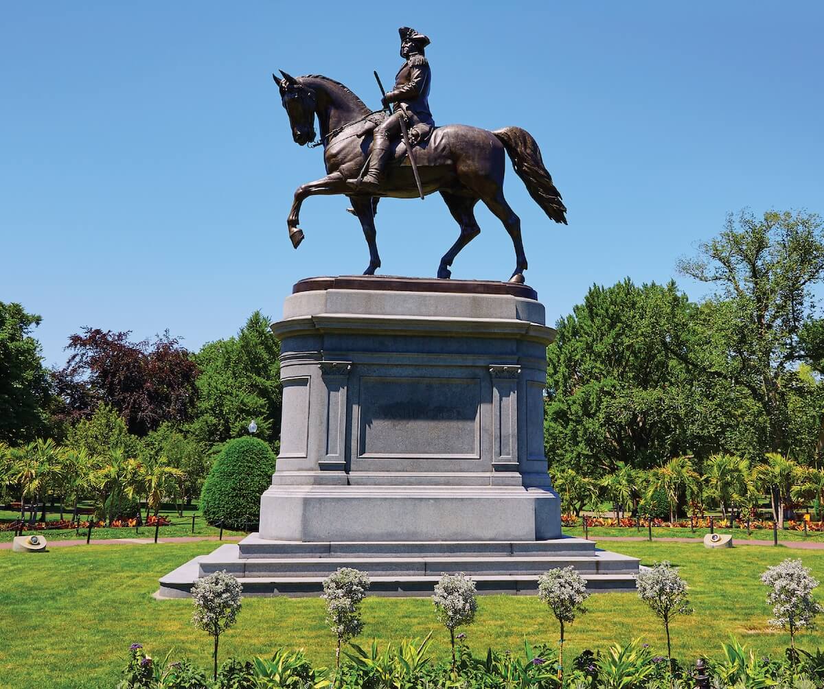 Bronze equestrian statue of George Washington in a tricorn hat, holding a baton and mounted on a walking horse atop a granite pedestal against a clear blue sky.