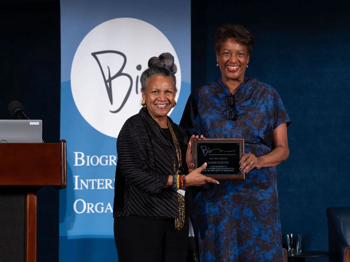 Two women stand on a stage smiling and holding a plaque together during an award presentation. Behind them is a blue backdrop with a “Bio” logo for Biographers International Organization, and a podium with a microphone is visible to the side. The plaque reads that the award is presented to Dawn Porter.