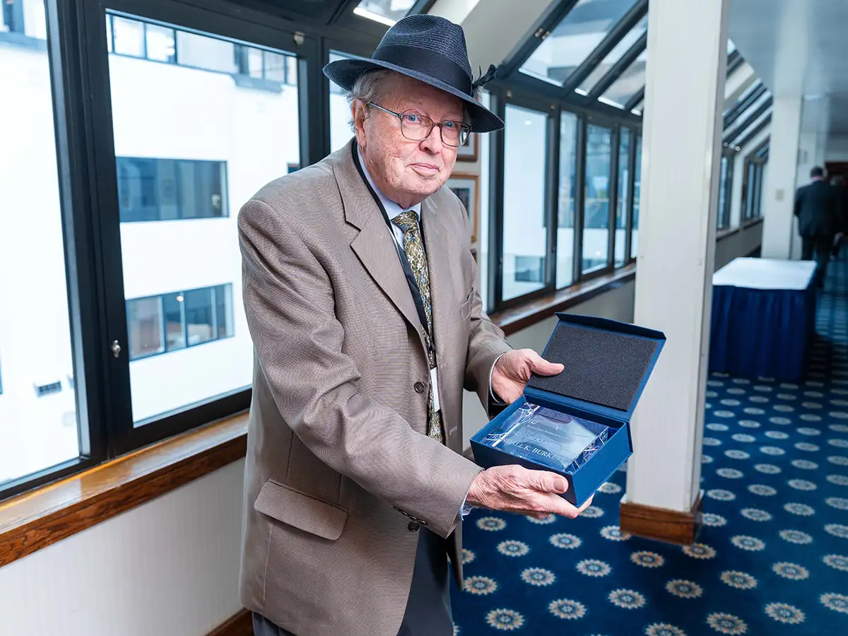 man wearing a tan jacket and blue fedora standing in an even hallway holding a box with an award
