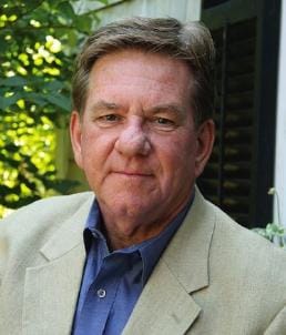 portrait of a man in a tan jacket and blue shirt standing outside in front of a building