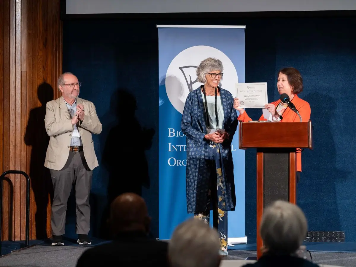 A woman at a podium presents a certificate to another woman standing beside her on a stage, while a man to the left applauds. The scene appears to be an award ceremony, with a banner in the background and an audience visible in the foreground.