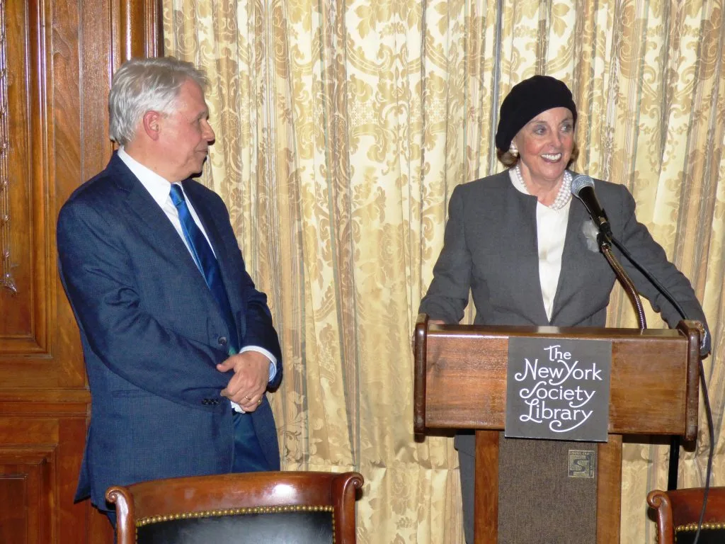 Woman speaking at podium at New York Society Library, man in suit looking on