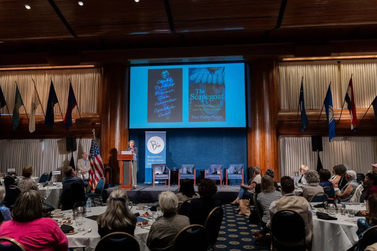 A speaker stands at a podium on a stage in a conference room, addressing an audience seated at round tables. Behind the speaker is a large screen displaying two book covers, including one titled “The Scapegoat.” Flags line the walls on both sides of the room, and several empty chairs are arranged on stage for a panel discussion. The audience is attentively facing the speaker.