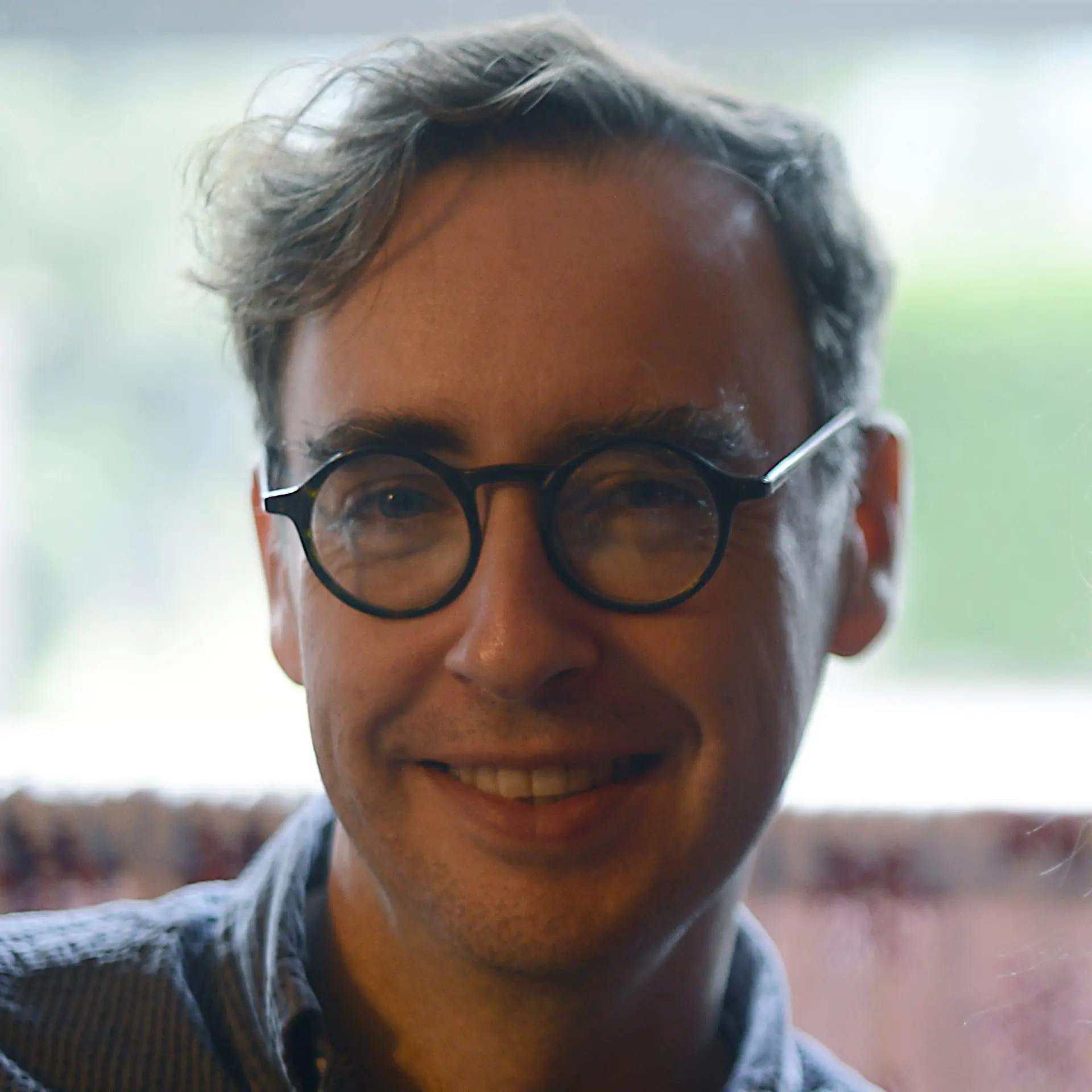 Close-up of smiling man with round black glasses and tousled hair indoors