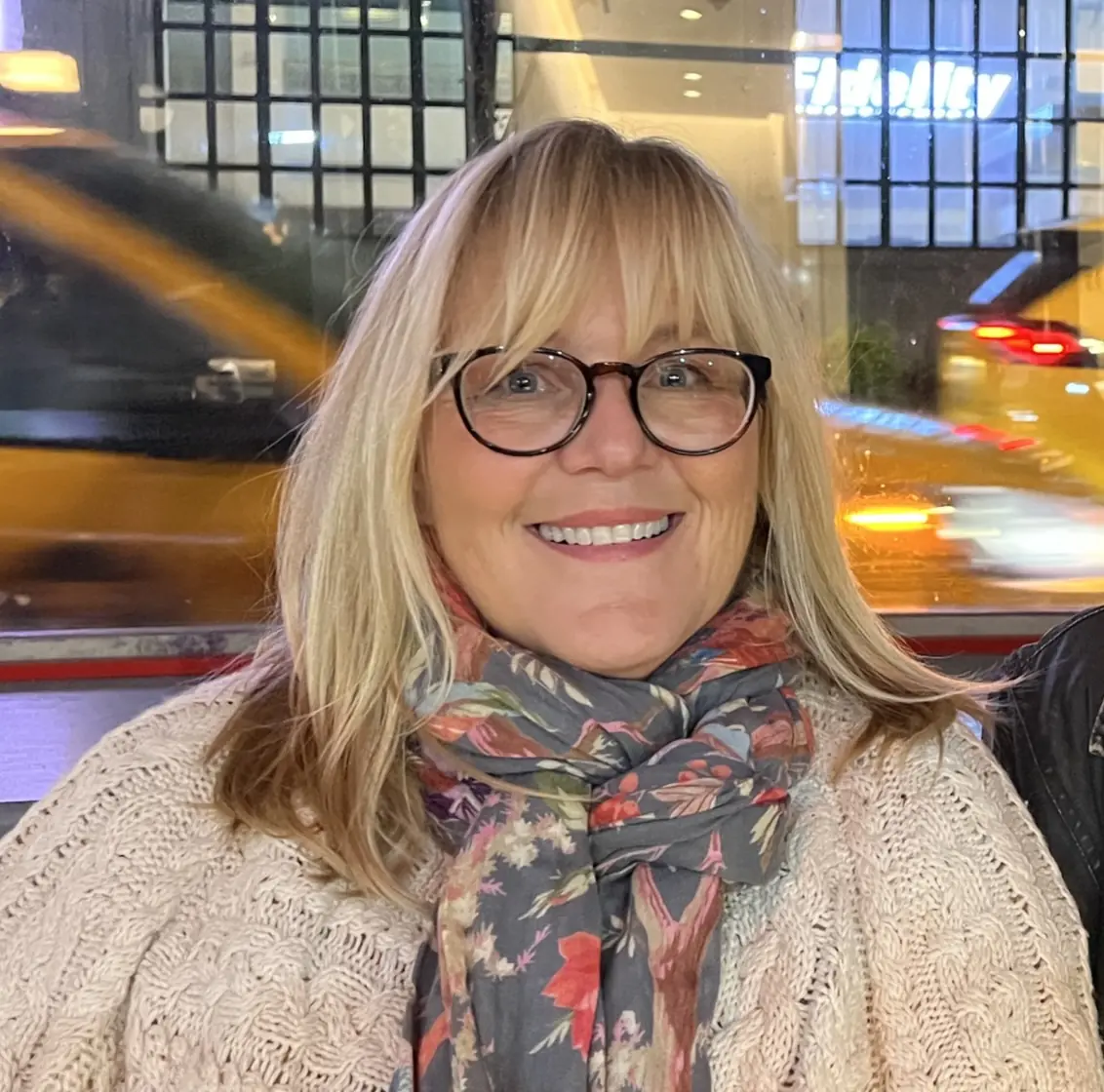 Smiling woman with blonde hair and glasses wearing floral scarf indoors at night
