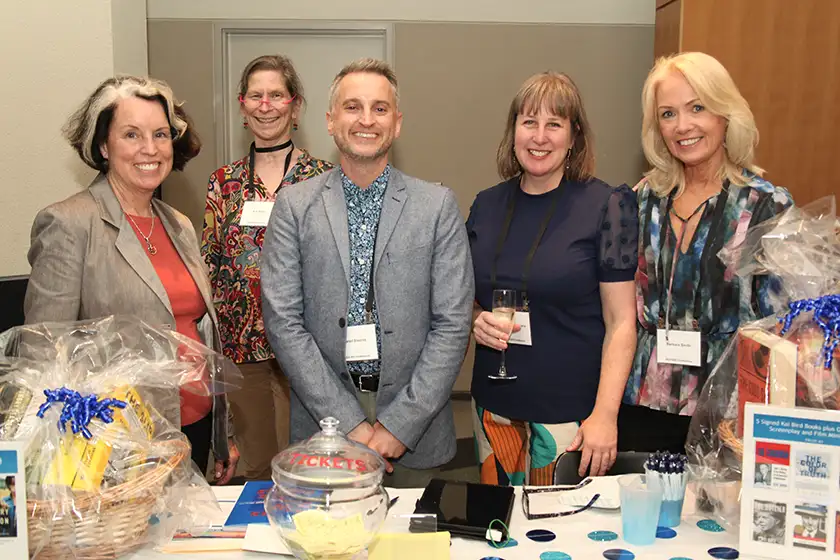 Five adults stand behind a table with gift baskets, raffle tickets, and event materials, smiling at the camera, in an indoor event setting.