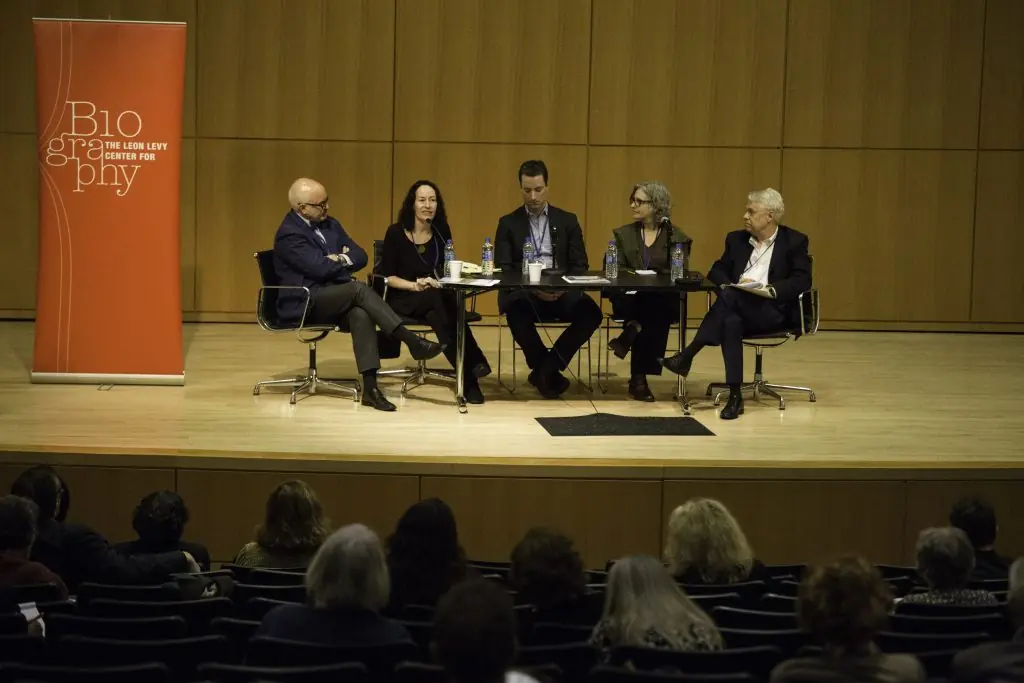 Five panelists with light skin tones sit at a table on a stage before an audience; a banner to the left reads "Biography THE LEON LEVY CENTER FOR".