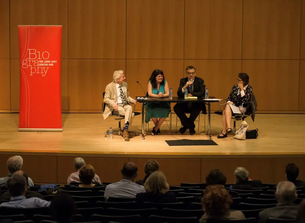 Four panelists with light skin tones sit at a long table on a stage; a red banner to the left reads "Biography THE LEON LEVY CENTER FOR".