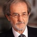 An individual with a light skin tone, grey hair, and a beard looks downward, wearing glasses, a dark suit, and a patterned tie.