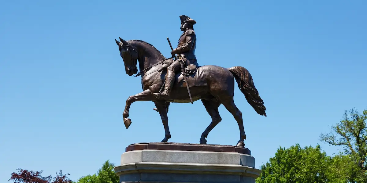 Bronze equestrian statue of George Washington in a tricorn hat, holding a baton and mounted on a walking horse atop a granite pedestal against a clear blue sky.