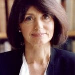 A person with a light skin tone and dark chin-length hair looks forward with a slight smile, wearing a dark blazer over a white top.