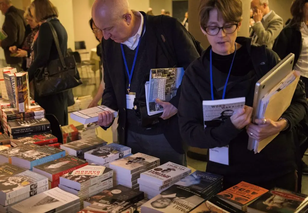 Two individuals with light skin tones look at books on a crowded display table; several titles including "WORD WARRIOR" and "DARROW" are visible.