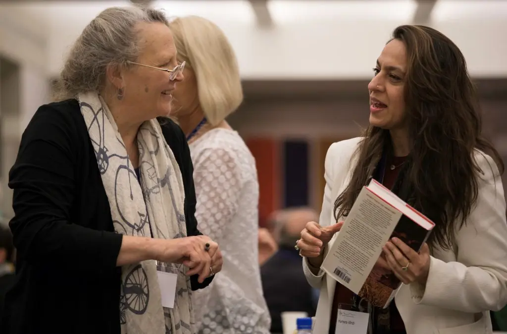 Two individuals, one with a light skin tone and grey hair and the other with a medium skin tone and long dark hair, converse while holding a book.
