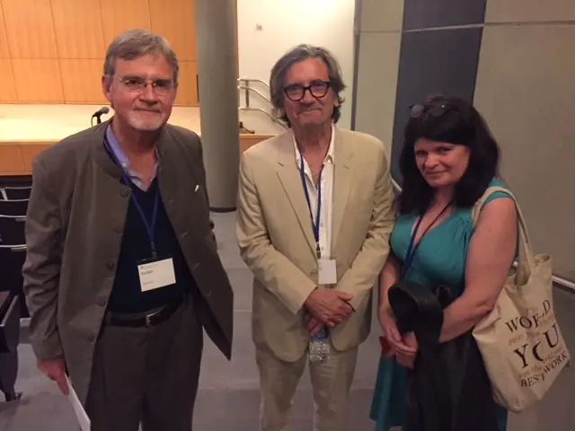 Three individuals with light skin tones stand together in an auditorium; two wear jackets and glasses, while one wears a teal dress.