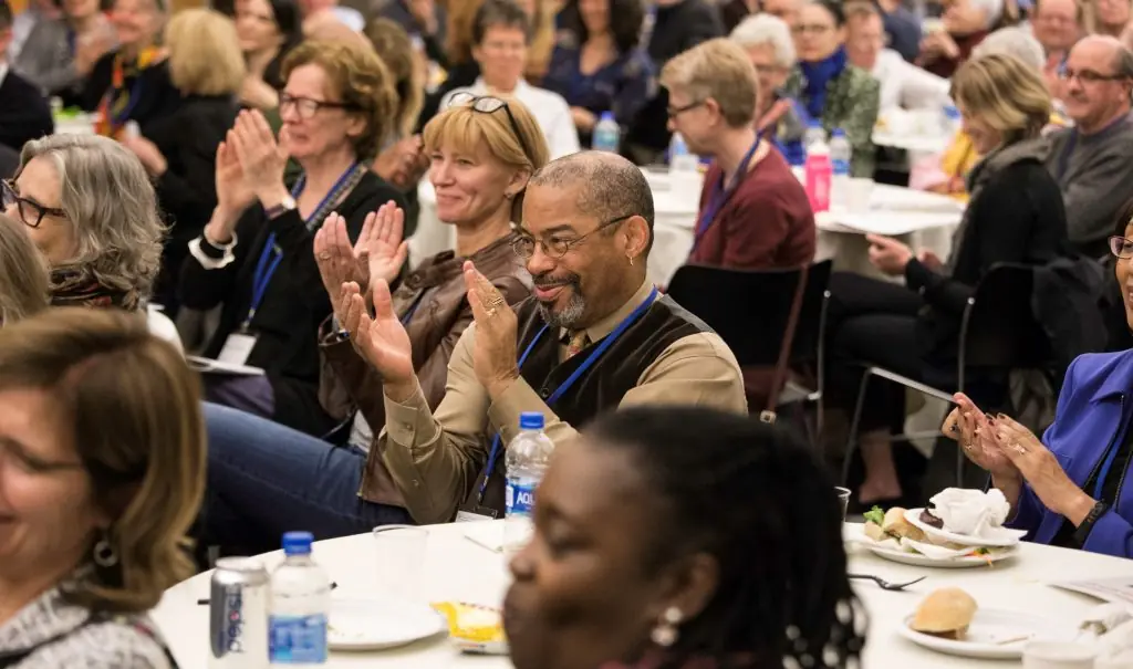 A diverse group of people sits at round tables, smiling and applauding during a luncheon event.