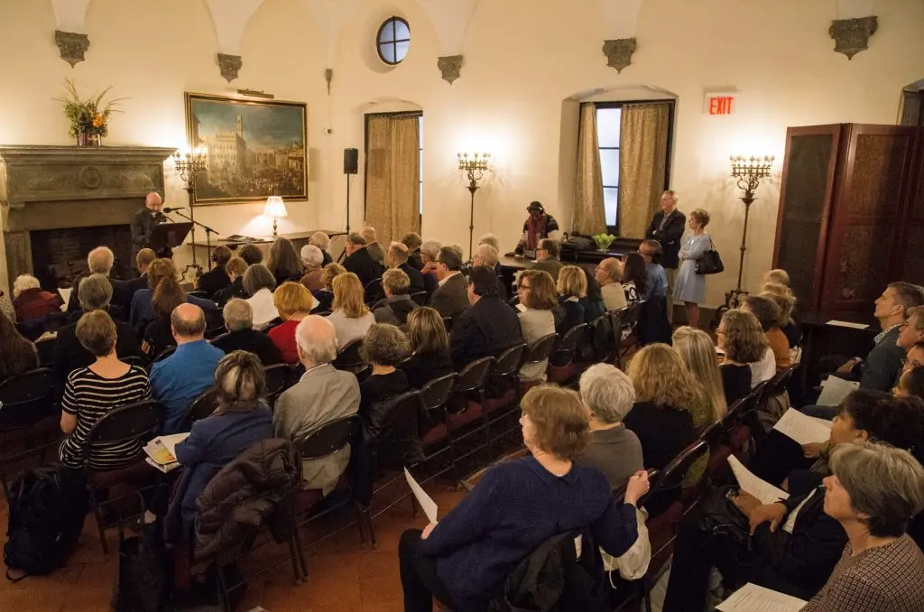 An audience sits in a high-ceilinged room facing an individual speaking at a podium near a large fireplace and a framed painting.