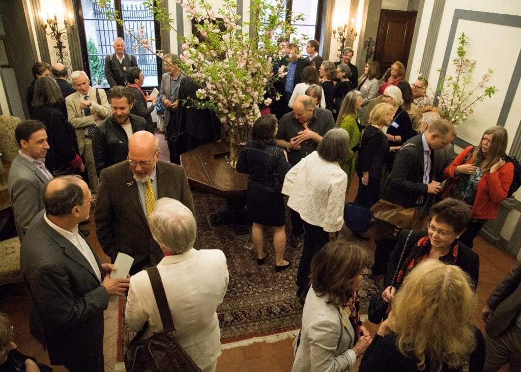 A large group of people with various skin tones stands and talks in an ornate room featuring a large floral centerpiece on a central table.