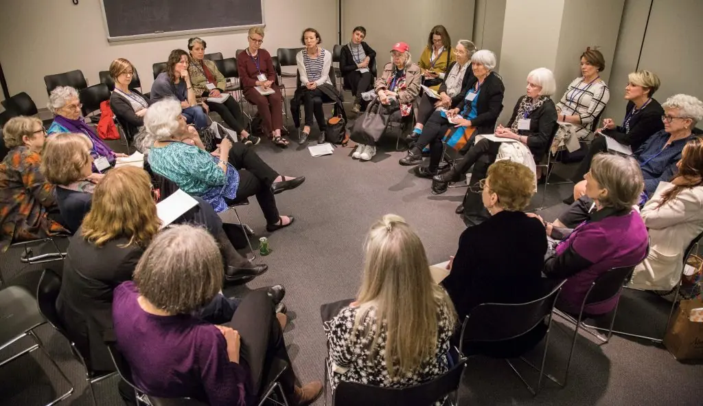 A large group of people with various skin tones sits in a circle of chairs for a discussion in a room with a chalkboard in the background.