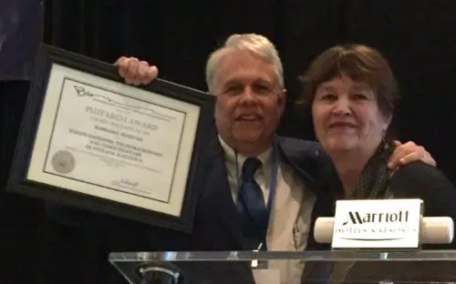 Two people with light skin tones smile together; one holds up a framed certificate titled "PLUTARCH AWARD," and a "Marriott" sign is visible.