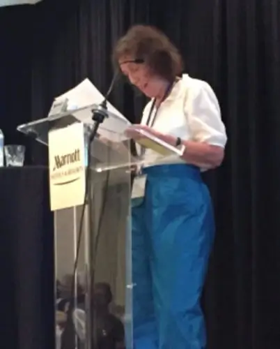 A person with a light skin tone stands at a Marriott podium, looking down at a book while speaking into a microphone.